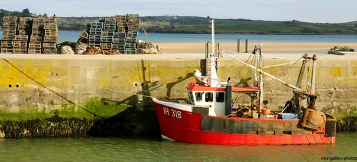 This landscape photograph shows a fishing boat docked in a harbour during the afternoon in winter in the United Kingdom. The boat, painted mainly red, features identification markings and equipment typical of fishing transport vessels and is moored beside a concrete quay. Stacks of lobster pots and ropes are visible on the harbour side, indicating the area's use for fishing activities. Behind the harbour wall, the scene extends to sandy banks and a tidal estuary, with green hills in the distance, characteristic of the British coastline. The image highlights boats as a vital form of transport within fishing communities along the United Kingdom's shores.